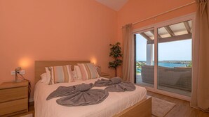 Ein Schlafzimmer mit einem großen Bett, einem Nachttisch, einer Topfpflanze und Blick auf das Meer durch eine Schiebetür.