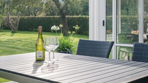 Mesa de patio con botella de vino y copas, rodeada de sillas de mimbre, con vista a un jardín y una puerta de cristal que da a un espacio al aire libre.