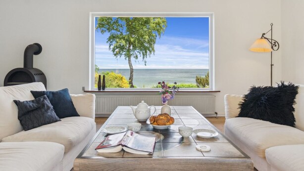 Un salon doté d’une grande fenêtre donnant sur un arbre et la mer, une table basse avec des livres et un vase de fleurs, et une lampe placée à droite.
