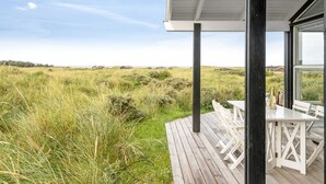 Une terrasse en bois avec des sièges extérieurs, offrant une vue sur un paysage verdoyant et des bâtiments situés au loin.
