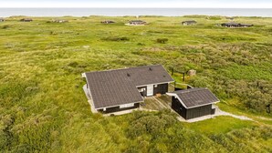 Une maison moderne dotée d’une grande terrasse et d’un garage séparé, entourée d’un champ verdoyant et bordée, au loin, par une étendue d’eau.
