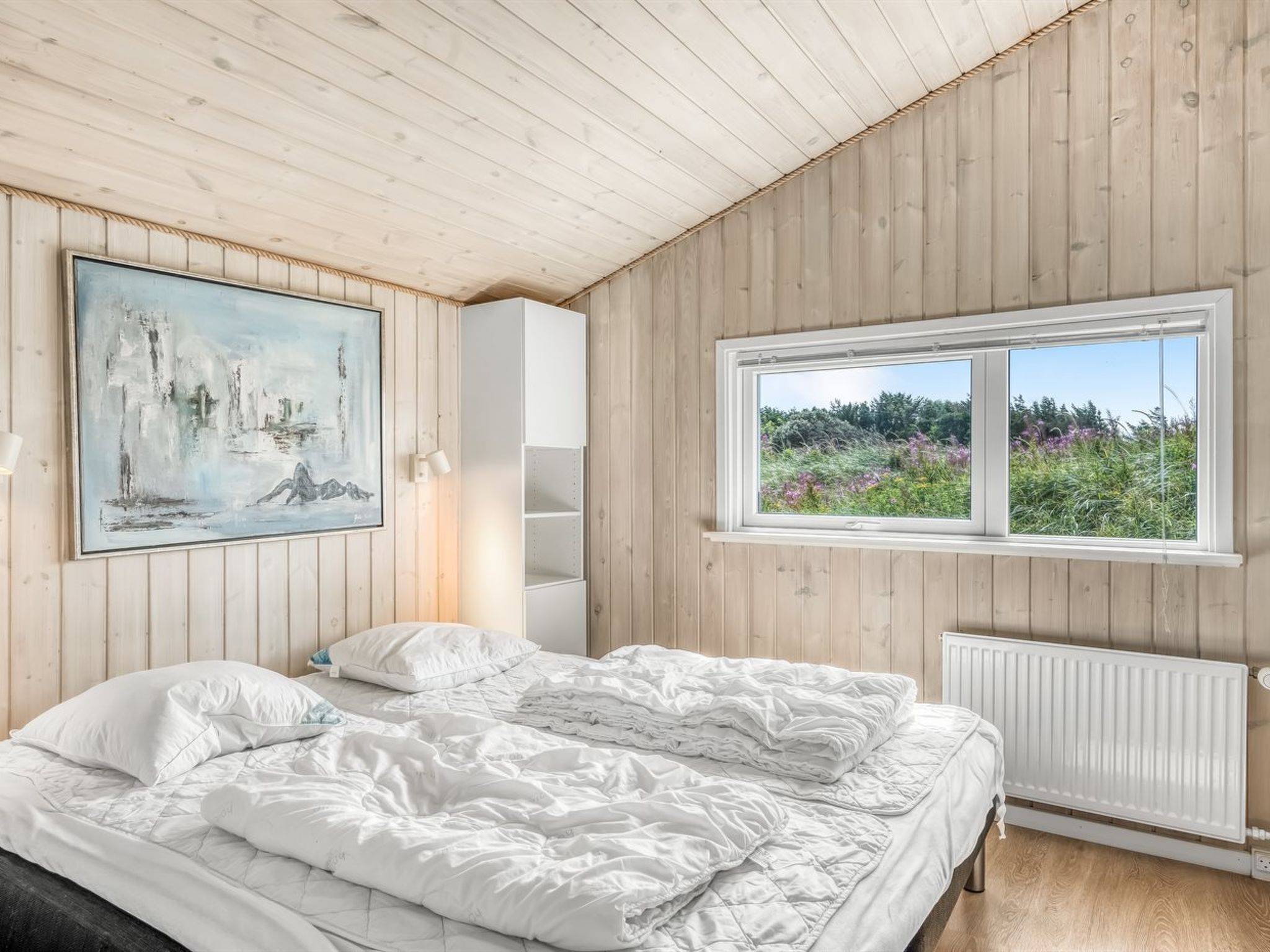 A bedroom with a bed, a painting, a window with a view of greenery, and a white radiator.