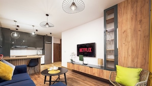 Una sala moderna con un sofá, una mesa de centro y una silla de mimbre. Un televisor está montado en la pared mostrando 