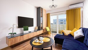 Una sala de estar moderna con un televisor de pantalla plana, un mueble de madera, una mesa redonda con juego de té, un sofá azul, cortinas amarillas y vistas de edificios a través de amplios ventanales.