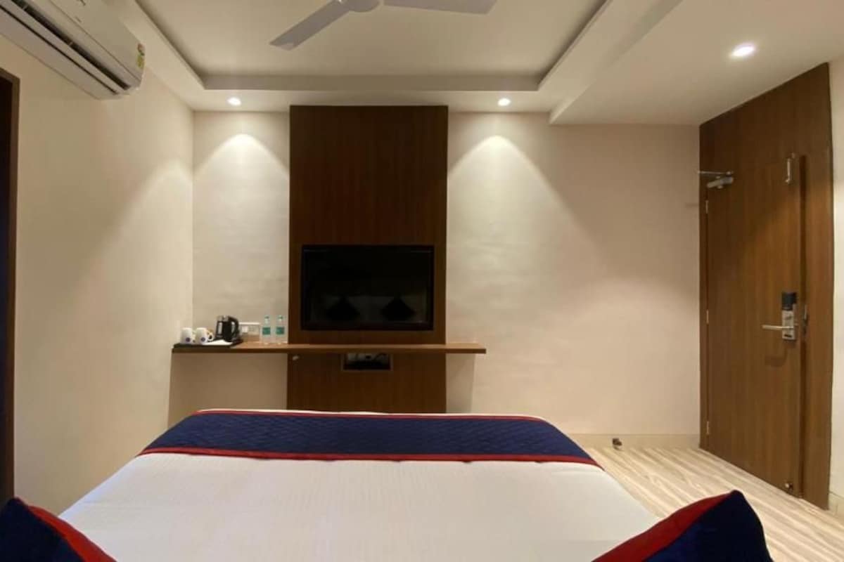 Habitación de hotel con cama, televisión instalada en la pared, armario de madera y baño visible a través de una puerta abierta.
