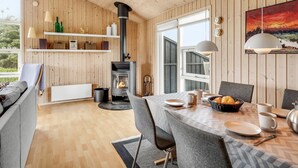 A cozy cabin interior with a wood-paneled wall, a central wood-burning stove, a dining table set for four, and a large window offering a view of the outdoors. - "Niesina" - 200m from the sea by Interhome (Løkken)