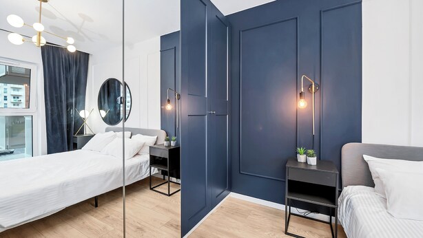A modern bedroom with two beds, a large mirror, bedside tables with lamps, and a view of a building through a window.