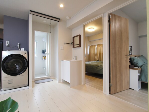 A compact, well-lit room with a washing machine, a small bathroom, and a bed.