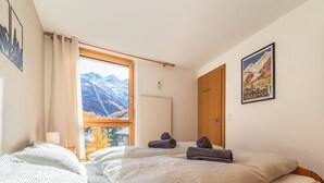 A bedroom with two beds, a window with a mountain view, a wooden door, and a poster on the wall. - Haus Judith - Bonita by Interhome (Saas-Fee)