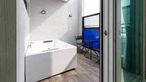 Un baño moderno con una bañera independiente, una mesita con una silla y una puerta de cristal que da a un balcón con barandilla azul.
