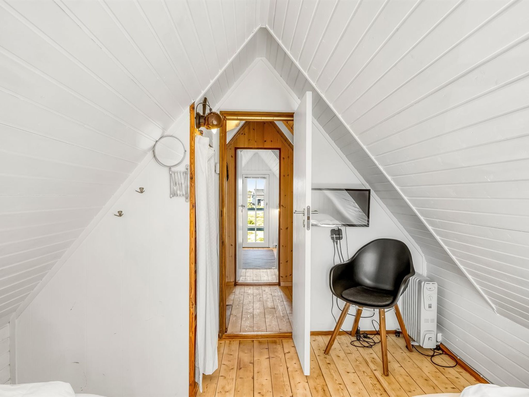 Ein schmales Schlafzimmer im Dachgeschoss mit Holztür zu einem Balkon, einem schwarzen Stuhl und einer Heizung.