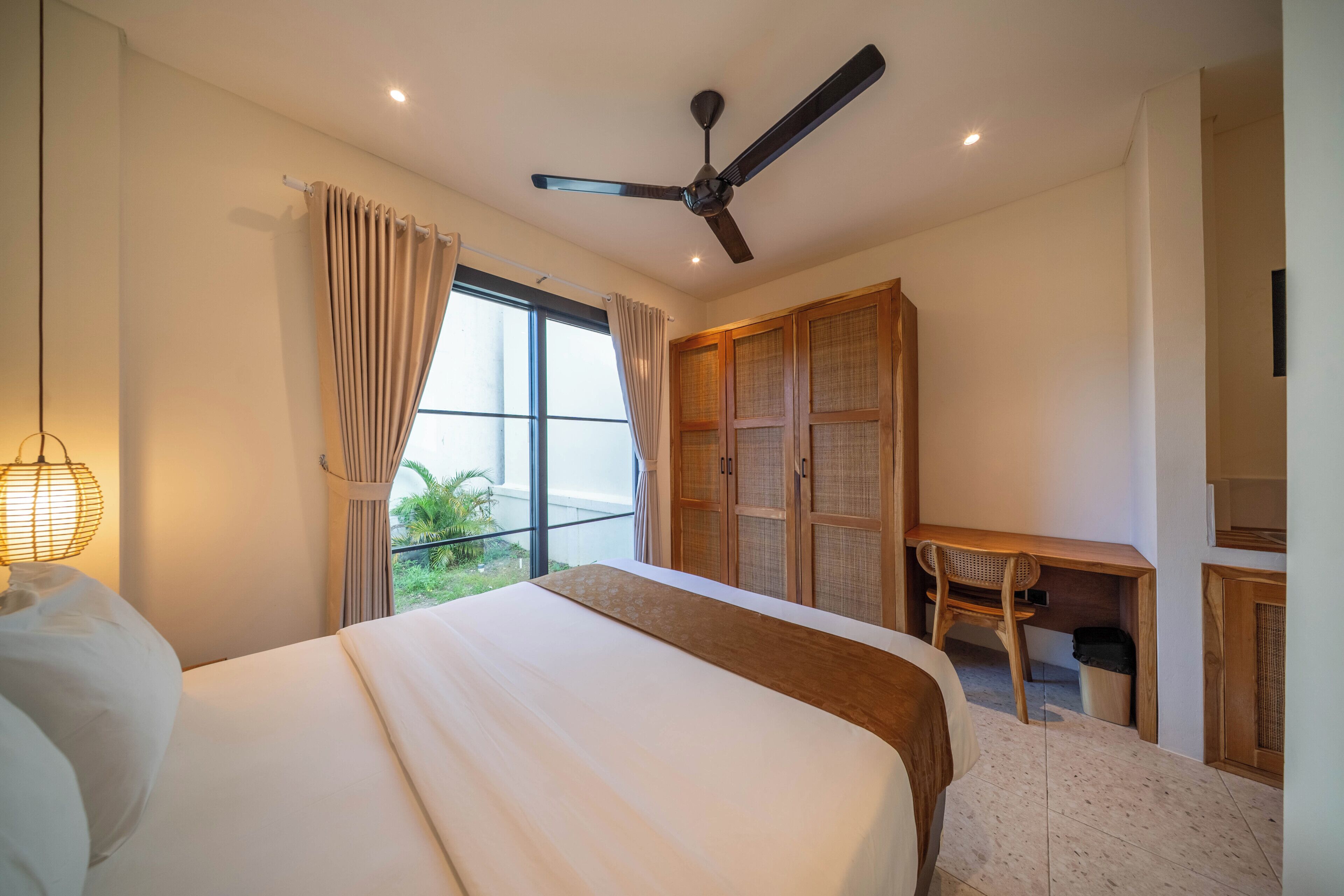 A bedroom with a bed, a desk, a chair, a ceiling fan, and a window with curtains.