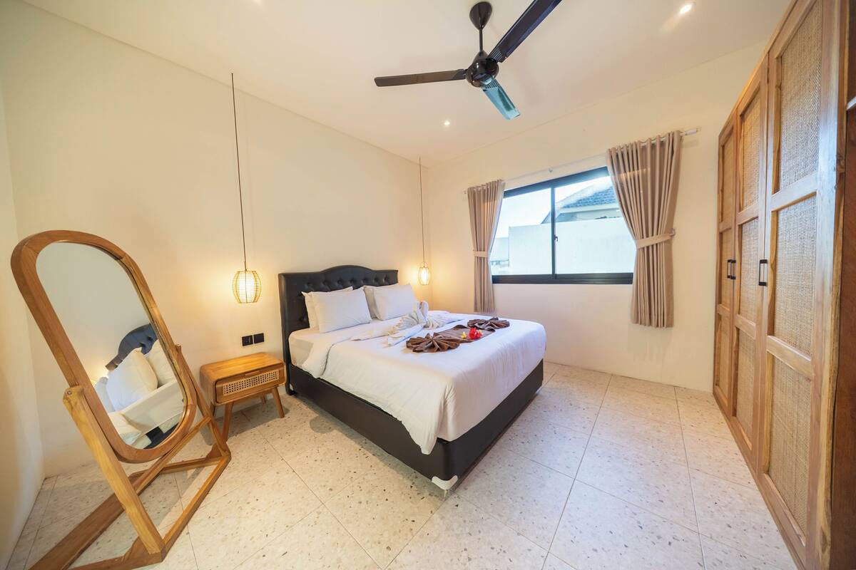 A bedroom with a bed, a ceiling fan, a window with curtains, a wooden wardrobe, and a mirror.
