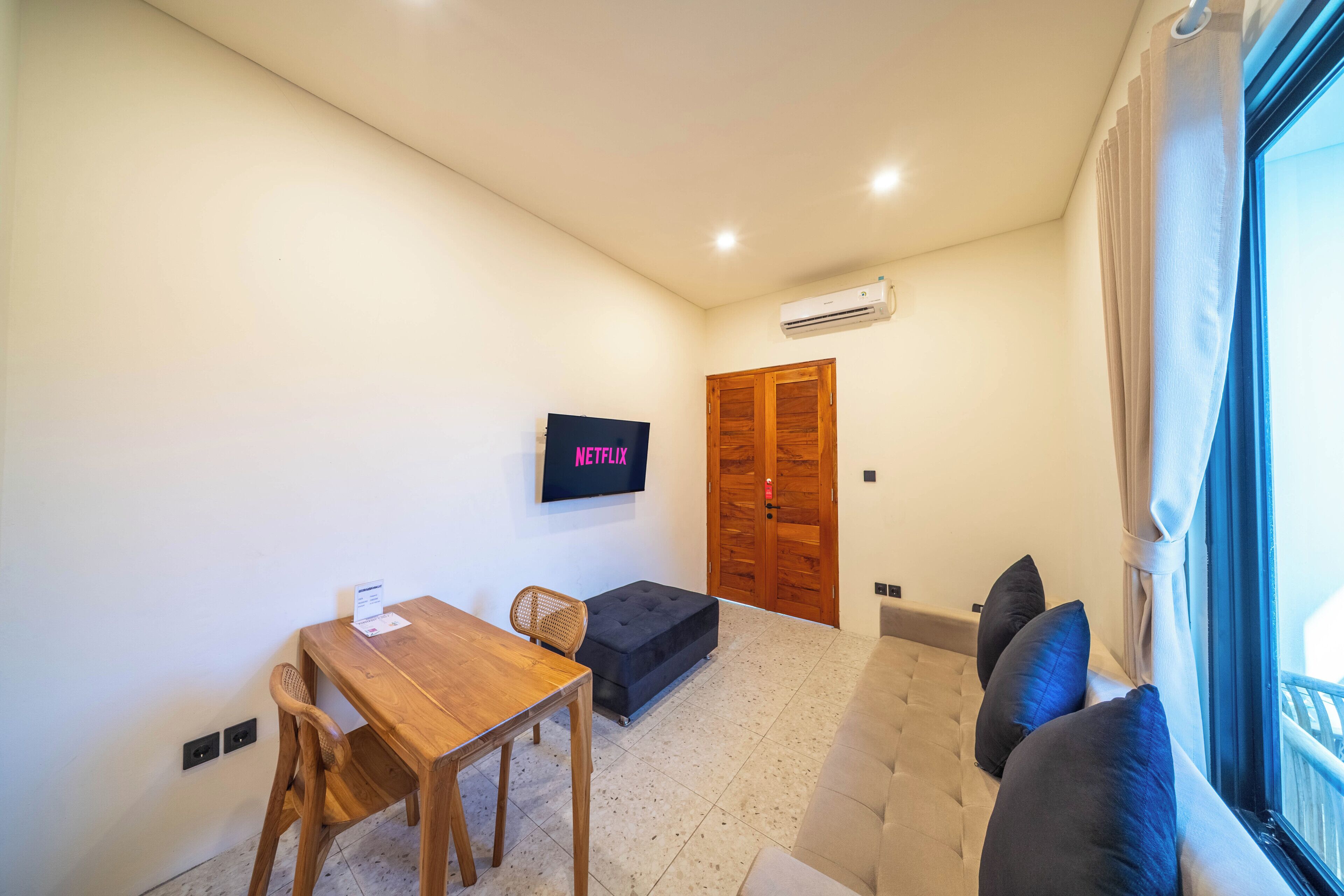 A living room with a wooden table, a sofa, a TV mounted on the wall, and a door.