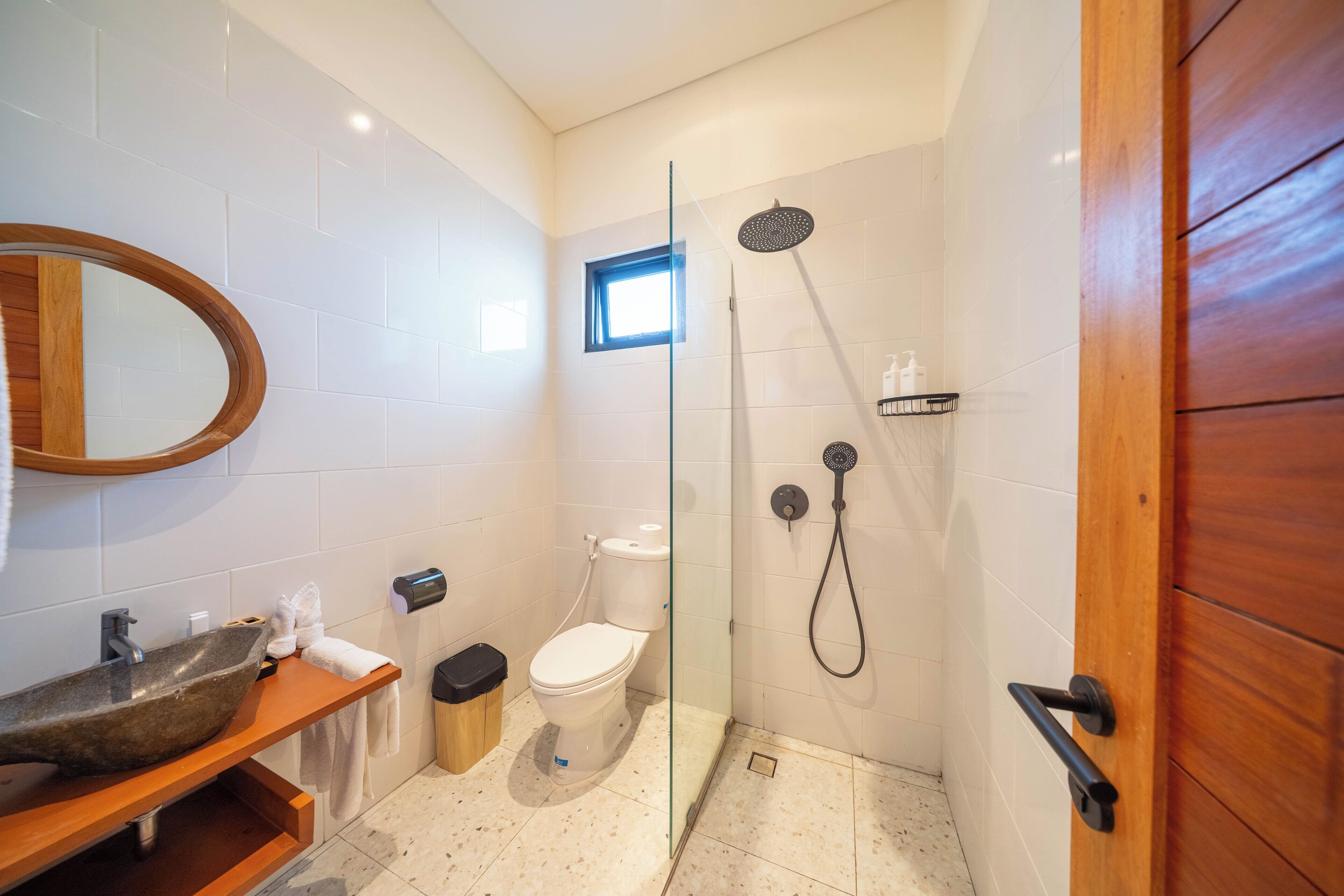 Et badeværelse med et trækommode, en glasbrusekabine, toilet og et lille vindue.