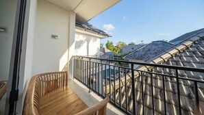 A balcony with a wooden bench, a metal railing, and a view of neighboring buildings and a swimming pool. - Lala Panzi Villa (Jimbaran)