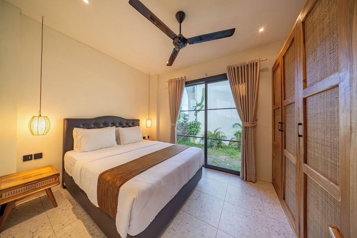 A bedroom with a bed, ceiling fan, bedside lamp, and a view of greenery through the window.