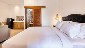A bedroom with a large bed, a wooden wardrobe, a kitchenette, and a door leading to another room. - Lala Panzi Villa (Jimbaran)