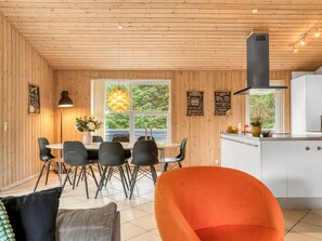 Un coin repas avec une table et des chaises, une fenêtre donnant sur un espace verdoyant, un coin cuisine avec une cuisinière et des placards, et une chaise orange vif.