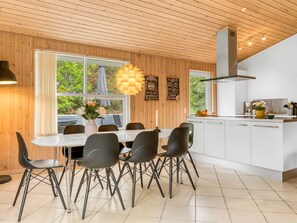 Una cocina moderna con armarios blancos, una zona de comedor con una mesa blanca y sillas negras, y un ventanal con vistas a zonas verdes.