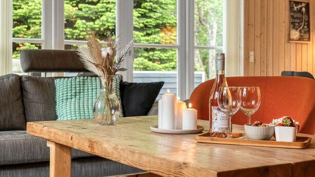 Ett matbord i trä med ljus, en vinflaska och glas på ett brickbord, placerat i ett rum med en grön, vadderad soffa och utsikt över träd genom stora fönster.