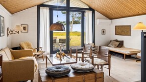 Ein gemütliches Zimmer mit schrägen Wänden, einem großen Fenster und Blick ins Freie. Es gibt ein Sofa, einen Sessel, einen Tisch mit Lampe und eine dekorative Truhe.