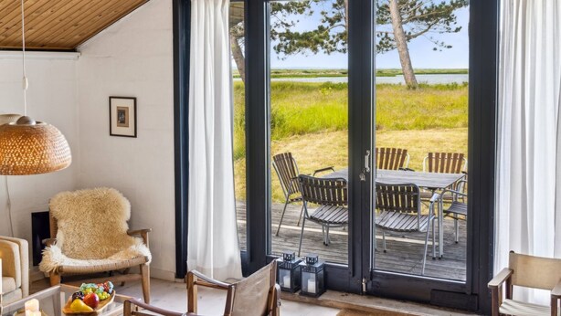 Ein gemütliches Zimmer mit Holzbalkendecke, einem großen Fenster mit Vorhängen, einem bequemen Sessel, einem Esstisch mit Stühlen und einem kleinen Tisch mit Kerzen und Früchten.