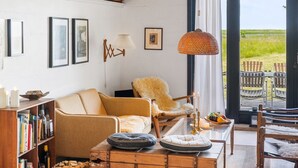 Ein gemütliches Wohnzimmer mit Holzbalkendecke, einer Couch, einem Sessel, einem Couchtisch und einem Bücherregal. Ein großes Fenster bietet einen Blick ins Freie.