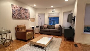 A modern living room with a wooden floor, a brown leather sofa, a white coffee table, and a grey sofa. There is a TV mounted on the wall, a floor lamp, and a large abstract painting on the wall. - Quiet, Cat-Enabled Paradise in the Heart of San Francisco (San Francisco)
