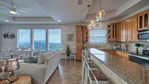 A spacious living area with a large sofa, dining table, and chairs. A kitchen with wooden cabinets and a granite countertop is visible. - Beach Boardwalk, Pool Access & Ocean Views | Coral Reef by AvantStay (Port Aransas)