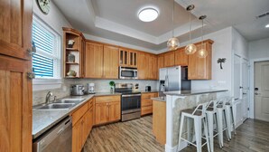 A modern kitchen with wooden cabinets, stainless steel appliances, a built-in oven, and a bar area with stools. - Beach Boardwalk, Pool Access & Ocean Views | Coral Reef by AvantStay (Port Aransas)