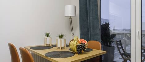 A dining area with a wooden table, four chairs, a fruit bowl, two potted plants, and a floor lamp.