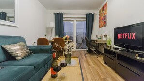 Un salon avec un canapé, une table basse sur laquelle se trouvent des verres à vin et une bouteille, un téléviseur affichant le logo « NETFLIX », et un coin repas avec une table et des chaises.