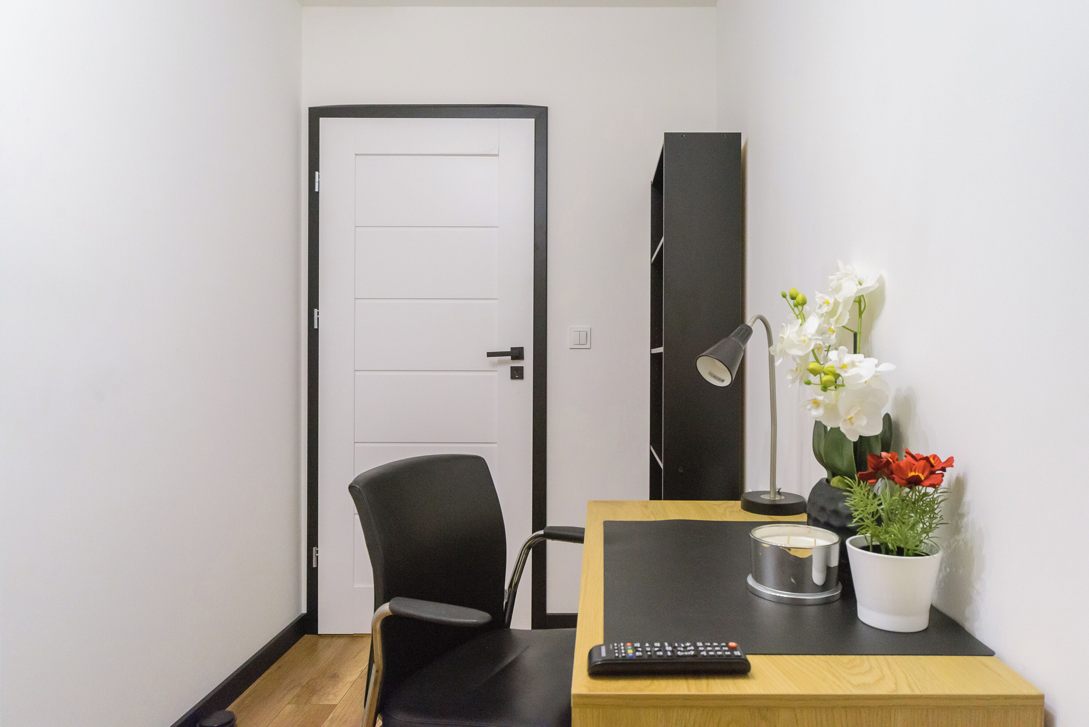 Een kamer met een houten bureau, een zwarte stoel, een afstandsbediening, een kaars en een bloeiende kamerplant.
