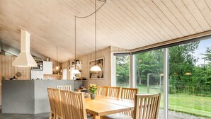 Een moderne keuken met houten lambrisering, een eethoek met een grote tafel en stoelen, en uitzicht op een weelderige tuin door grote ramen.