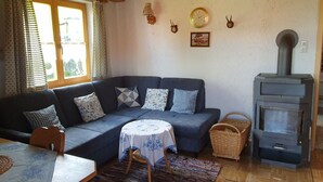 A cozy living room with a blue sofa, a round table with a floral tablecloth, a woven basket, and a wood-burning stove. - Ferienhaus für 5 Personen, 2 Seperate Schlafzimmer (Anger)