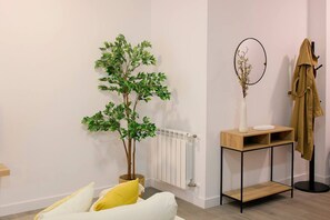Una habitación con un sofá blanco, una planta pequeña, un radiador, una consola de madera y un abrigo colgado en un gancho.