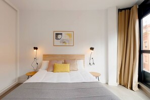 A hotel room with a bed, two bedside tables, a headboard, a wall-mounted light fixture, a window with curtains, and a framed picture on the wall.