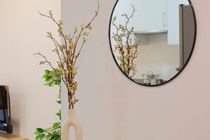 Una habitación con un espejo redondo en la pared, un jarrón blanco con ramas y flores amarillas, y un televisor sobre un soporte.