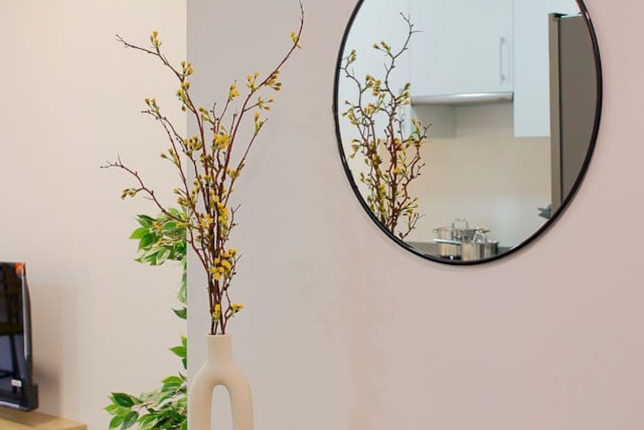Une pièce avec un grand miroir rond au mur, un vase blanc orné de branches et de fleurs jaunes, et un téléviseur posé sur un meuble.