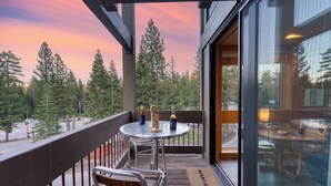 Un balcon avec vue sur les arbres et un coucher de soleil. On y trouve une table avec des bouteilles et un jeu de société.