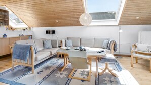 Um loft espaçoso com pé-alto, um claraboia, uma área de estar aconchegante com sofá e poltronas, uma mesa de jantar e uma cozinha com armários e uma janela com vista para o quintal.