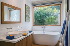 Ein modernes Badezimmer mit freistehender Badewanne, zwei Waschbecken, einem großen Spiegel und einem Fenster mit Blick auf Grünflächen.