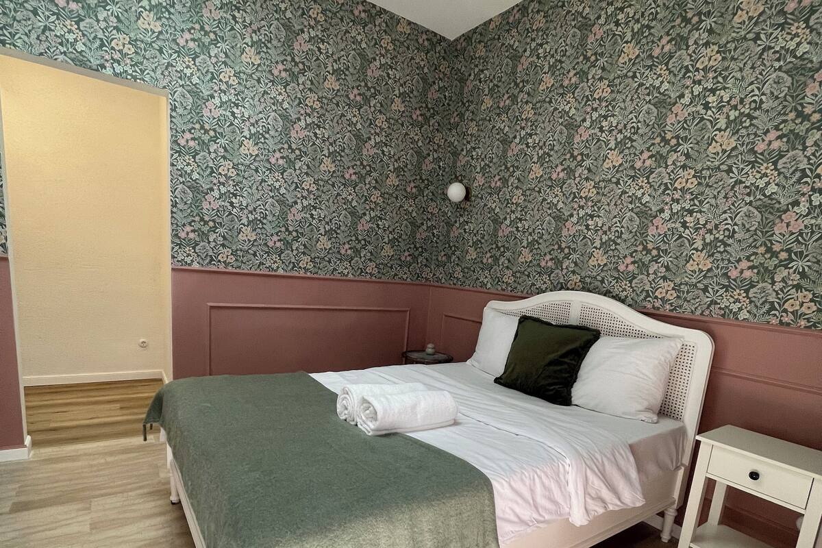 A bedroom with a bed, a bedside table, a wall with floral wallpaper, and a skylight.