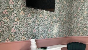 Une pièce comprenant une console blanche, une chaise verte, un panier en osier et un téléviseur fixé au mur.