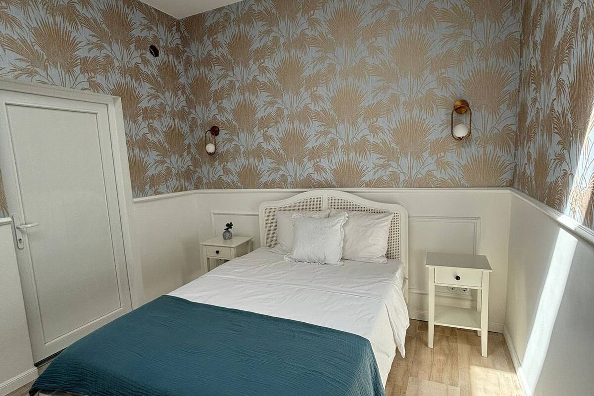 A bedroom with a bed, white nightstands, a wardrobe, and patterned wallpaper.