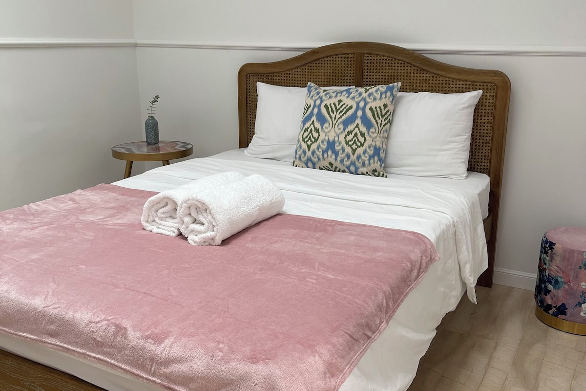 A neatly made bed with a pink blanket and white linens, a bedside table with a vase, and a wall-mounted light fixture.