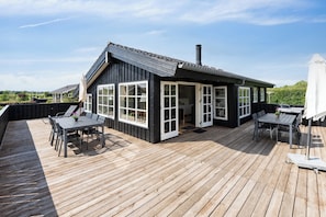 Eine Holzdachterrasse mit Essbereich, überdachter Lounge-Ecke und einem Gebäude mit großen Fenstern und Schornstein.