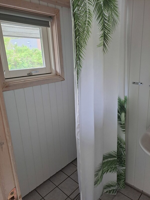 A bathroom with a window, a shower curtain with a palm leaf pattern, and a tiled floor. - Cozy summer house and annex in tranquility (Glesborg)