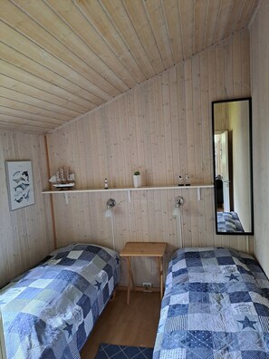 A room with wooden panel walls, two beds with blue and white patterned bedding, a small wooden table, a shelf with decorative items, and a mirror.